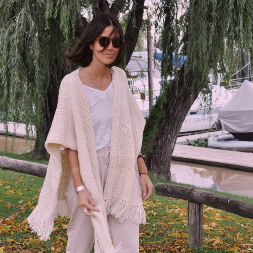 Woman in a white outfit with a beige cardigan standing on a leaf-covered ground with trees and water in the background.